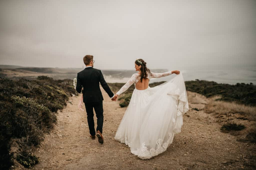 a bride and groom walking down a dirt road with the ocean behind them