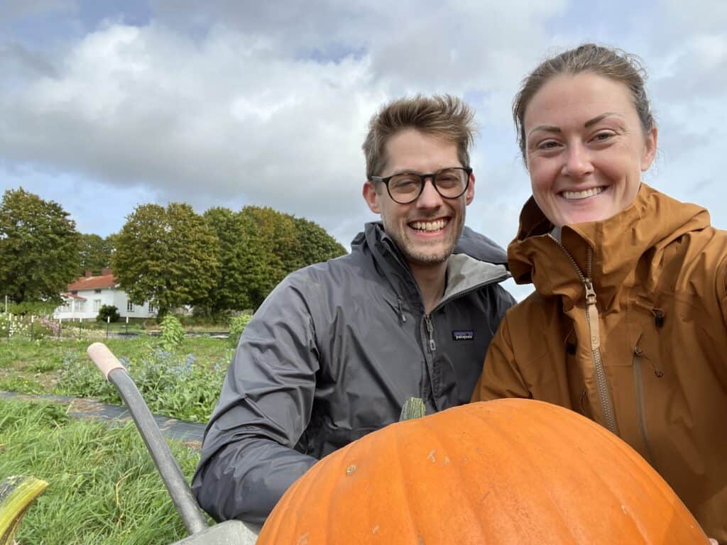 a couple smiling with a pumpkin and flower farm and house in the background